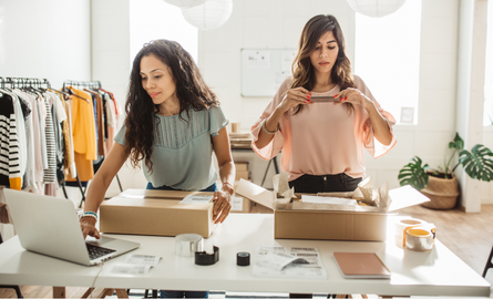two women standing at a desk with boxes and a computer